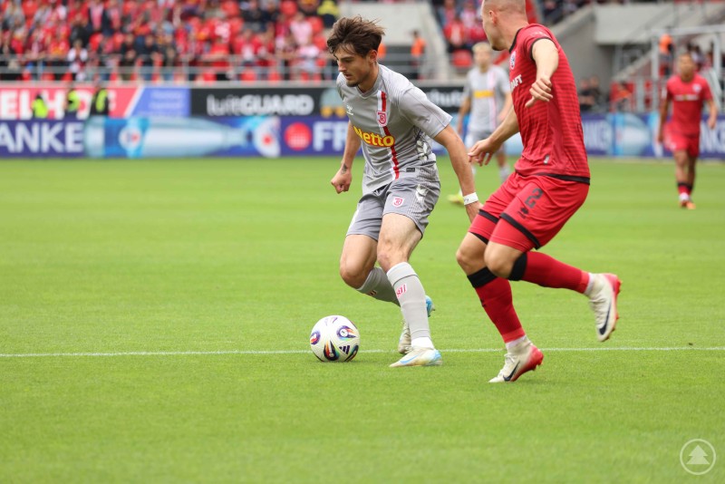 Spieler des SSV Jahn Regensburg am Ball, bedrängt von einem Gegenspieler des FC Ingolstadt, mit gefüllter Tribüne im Hintergrund.