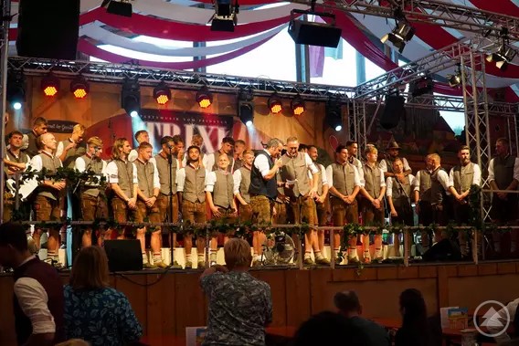 Auf dem ersten Bild ist eine große Gruppe von Männern und Frauen in Tracht auf einer Bühne zu sehen. Sie tragen Lederhosen, Westen und weiße Hemden, während im Hintergrund eine festlich geschmückte Halle mit Lichtern und roten Stoffbahnen zu erkennen ist.