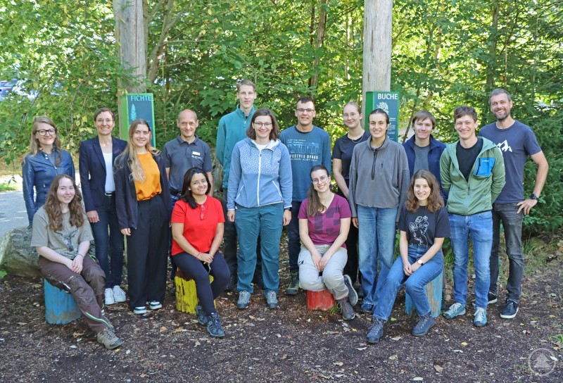Die neuen Freiwilligen mit Nationalparkleiterin Ursula Schuster und ihren Betreuern. Eine Gruppe von jungen Erwachsenen und Betreuern steht im Grünen, sie lächeln für ein Gruppenfoto vor Schildern mit Baumarten.