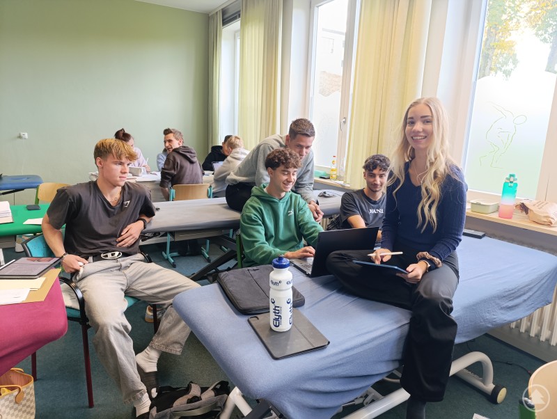 Gute Laune im Klassenraum – die angehenden Physiotherapeutinnen und Physiotherapeuten lernen im Team und tauschen sich rege aus. Gruppe von Schülerinnen und Schülern sitzt an Behandlungsliegen mit Laptops und Tablets und lacht.