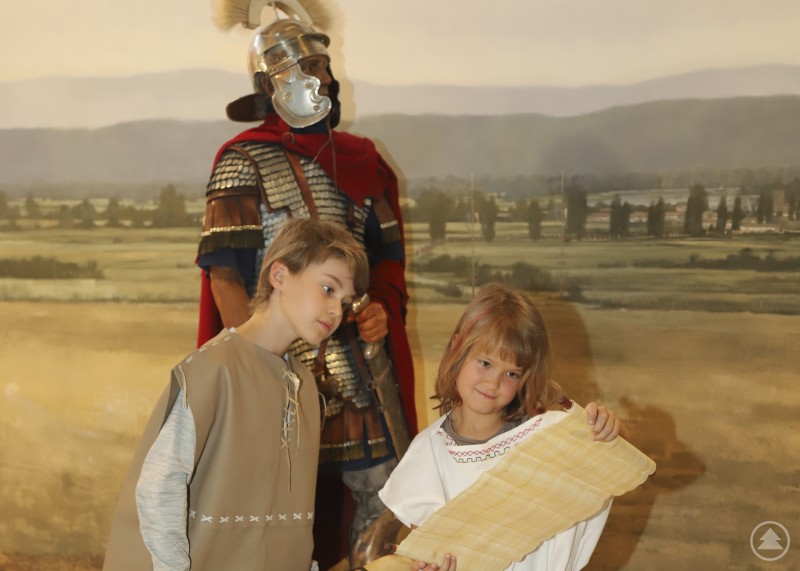 Spielerisches Lernen gehört im Museum Quintana fest zum museumspädagogischen Konzept. Zwei Kinder stehen vor einer Kulisse mit römischem Soldaten und halten eine Schriftrolle, die an ein antikes Dokument erinnert.