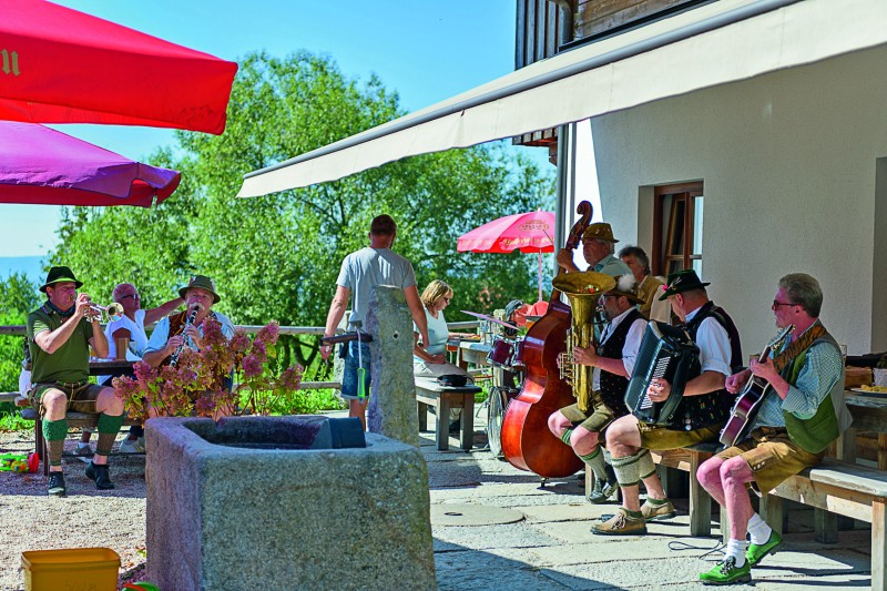 Zünftig aufgespielt wird am 18. Juni in den Bodenmaiser Biergärten. Eine bayerische Volksmusikgruppe spielt im Biergarten bei sonnigem Wetter. Gäste sitzen an Tischen unter bunten Sonnenschirmen, während Musiker in Tracht mit Tuba, Akkordeon, Trompete und Gitarre traditionelle Musik machen.