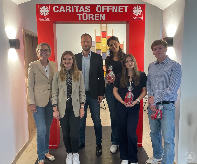 Freude über den Ausbildungsstart: Caritasvorstand Andrea Anderlik (v.l.), Ausbildungsleiterin Sabrina Hoffmann, Personalabteilungsleiter Thomas Furthmeier und die neuen Auszubildenden Mina Rakic, Anna-Lena Voggenreiter und Nico Fahl. Eine Gruppe von sechs Personen steht vor einem roten Rahmen mit der Aufschrift „Caritas öffnet Türen“. Drei der jungen Menschen halten kleine Geschenke in der Hand.