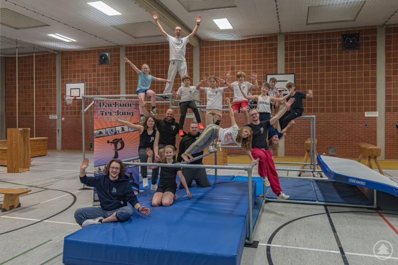 Die neuen Traceure trainieren gemeinsam mit den erfahrenen Parkour Läufern. Eine Gruppe junger Parkour-Sportlerinnen und -Sportler posiert in einer Turnhalle auf Matten und einem Stangengerüst. Einige stehen, sitzen oder balancieren auf verschiedenen Elementen und zeigen dynamische Posen.