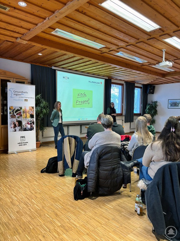 Zwei Referentinnen stehen vor einer Projektionsfläche und beantworten Fragen der Teilnehmenden. Das Publikum sitzt in mehreren Reihen, einige Jacken und Getränkeflaschen liegen am Boden.