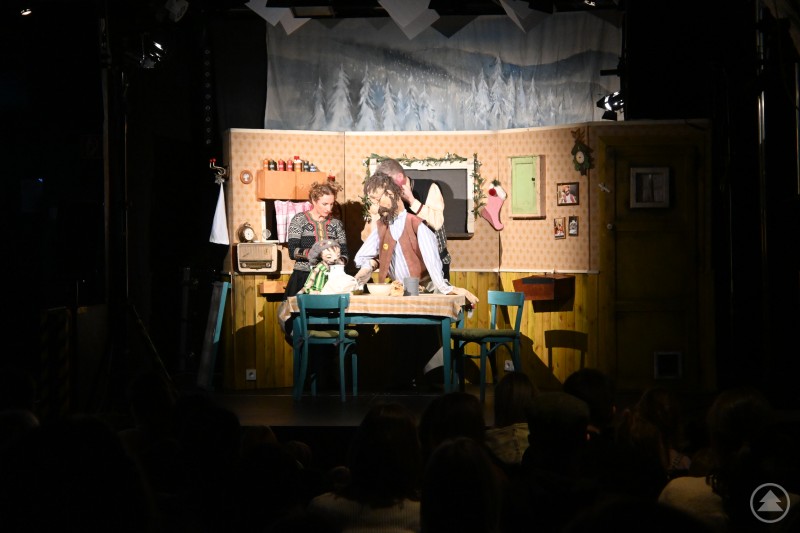 Szene aus dem Stück „Pettersson kriegt Weihnachtsbesuch“ Eine stimmungsvolle Szene aus dem Kindertheater zeigt die Figuren Pettersson, Findus und weitere Charaktere an einem gedeckten Tisch in einer liebevoll gestalteten Stube. Das Bühnenbild vermittelt eine winterliche, gemütliche Atmosphäre mit weihnachtlichen Dekorationen, während die Puppen- und Schauspielkunst im Mittelpunkt steht.