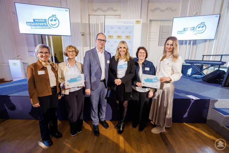 Zum Gruppenbild des ausgezeichnet guten Tons stellten sich zum Beispiel hier zu Staatsministerin Anna Stolz und MdL Martin Behringer die Rektorin Astrid Glaser v.l. mit Stellvertreterin Anneliese Stadler aus Hinterschmiding-Grainet und aus Thurmansbang-Preyung Lehrerin Andrea Freudenauer v.r. sowie Rektorin Renate Wilhelm. Mehrere Personen stehen auf einer Bühne und halten Urkunden in den Händen, im Hintergrund sind Bildschirme mit dem Logo Musikbegeisterte Grundschule zu sehen.