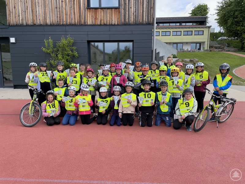 Die stolzen Viertklässler der Heinz-Teuerjahr-Schule Neuschönau präsentieren nach bestandener Radfahrprüfung ihre Urkunden. Gruppe von etwa 30 Grundschulkindern in Warnwesten und Fahrradhelmen posiert mit Urkunden vor einem modernen Schulgebäude. Zwei Fahrräder sind ebenfalls zu sehen.