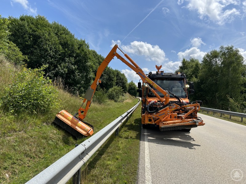 Seit Mai wird entlang der Kreisstraßen wieder gemäht, dieses Jahr aber zum Teil nach einem neuen Schema. in orangefarbenes Straßenunterhaltsfahrzeug mit Mäharm pflegt den Straßenrand entlang einer Kreisstraße, im Hintergrund dichter Wald und blauer Himmel mit Wolken.