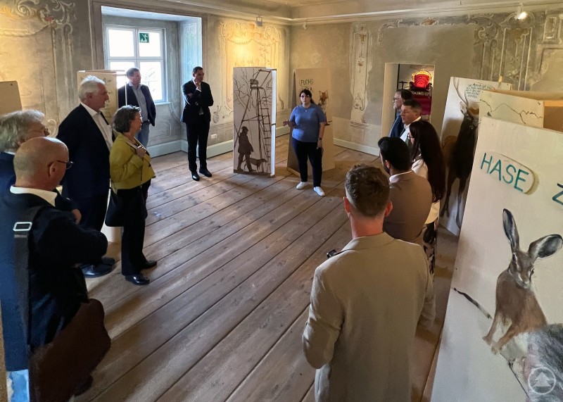 Besucherinnen und Besucher des BBA-Präsidentengesprächs im Schloss Wolfstein lauschen den Erläuterungen von Kulturreferentin Gabriela Rauscher. Eine Gruppe von Menschen steht in einem historischen Museumsraum, während eine Frau in der Mitte eine Führung hält.