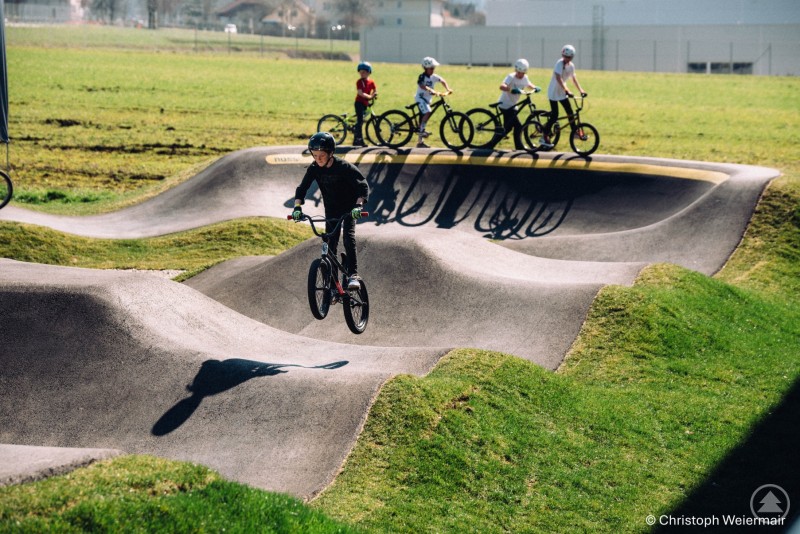 BMX-Fahrer nutzen die Wellen und Kurven des Pumptracks sportlich und mit viel Schwung. Jugendliche mit BMX-Rädern fahren über einen Pumptrack, einer springt gerade über eine Welle, im Hintergrund warten weitere Fahrer.