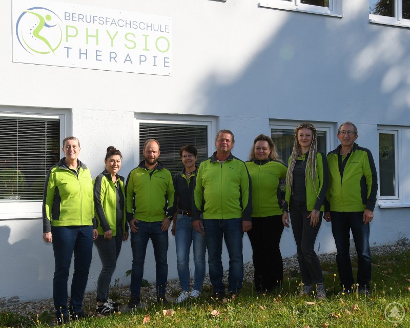Acht Personen stehen nebeneinander vor einer weißen Wand mit dem Schild „Berufsfachschule für Physiotherapie“.