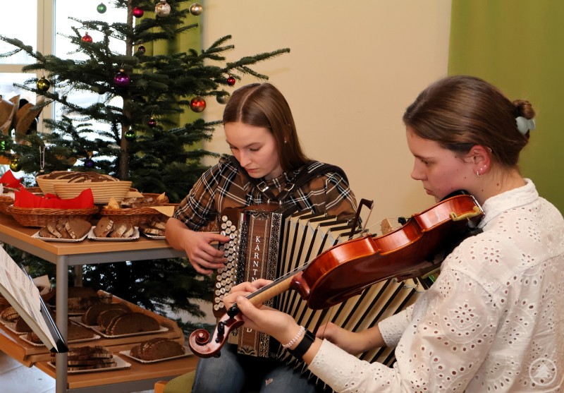 Musikalisch umrahmt wurde die Feierstunde von den Studierenden Johanna Stadler (rechts) und Lorena Wimmer (links) an Geige und Akkordeon. Zwei junge Frauen musizieren bei der Weihnachtsfeier. Eine spielt Geige, die andere Akkordeon. Im Hintergrund stehen ein geschmückter Weihnachtsbaum sowie ein Tisch mit Brot und Gebäck.