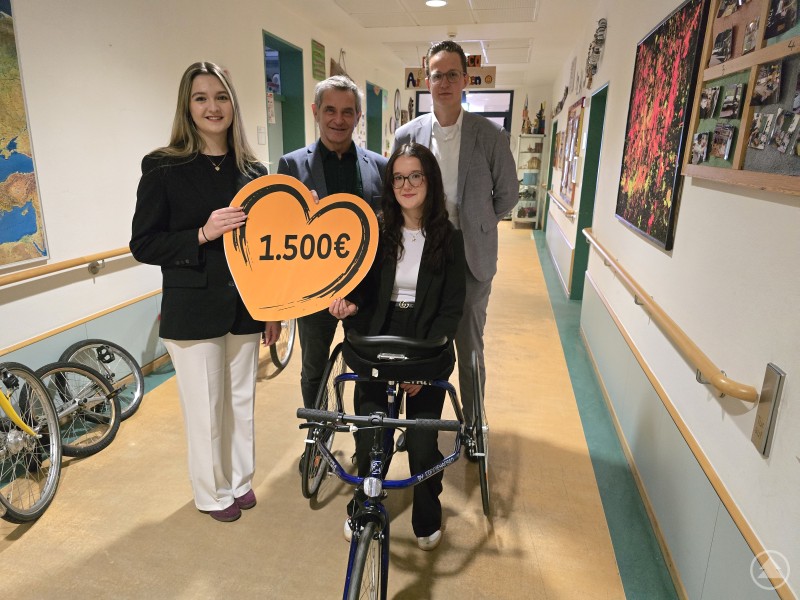 Die Azubis Anna Hofmann li. und Elena Kneidinger bringen mit 1500 Euro richtig Bewegung nach Don Bosco. Über das Sport-Bike freut sich Karl Bischof mit Alexander Stumpf. Zwei junge Frauen halten ein großes Herz mit der Aufschrift 1.500 Euro, daneben stehen Vertreter der VR-Bank und des Caritaszentrums Don Bosco. Vor ihnen ist ein blaues Sport-Bike für Jugendliche mit Handicap zu sehen.