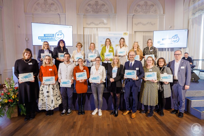 Zum Gruppenbild des ausgezeichnet guten Tons stellten sich zum Beispiel hier zu Staatsministerin Anna Stolz und MdL Martin Behringer die Rektorin Astrid Glaser v.l. mit Stellvertreterin Anneliese Stadler aus Hinterschmiding-Grainet und aus Thurmansbang-Preyung Lehrerin Andrea Freudenauer v.r. sowie Rektorin Renate Wilhelm. Große Gruppe von ausgezeichneten Lehrkräften und Verantwortlichen steht gemeinsam auf einer Bühne und präsentiert Urkunden für musikbegeisterte Grundschulen.