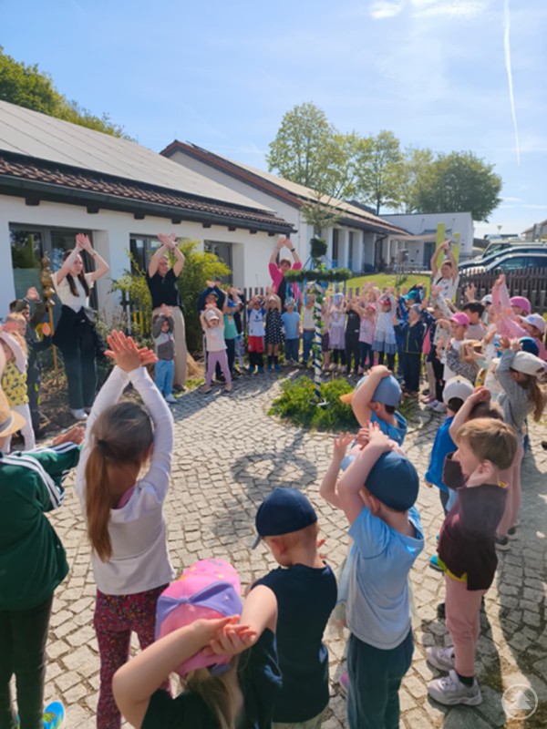 Kinder und Betreuerinnen tanzen gemeinsam rund um einen bunt geschm&uuml;ckten Maibaum auf dem Gel&auml;nde des Kindergartens.