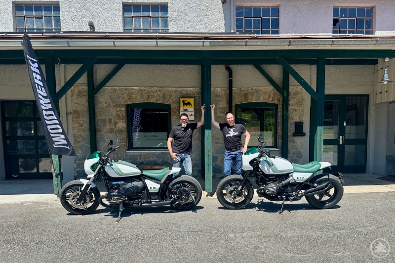 Ralf Eggl (rechts) und sein Teamkollege präsentieren die „Speed Sisters“ vor der Werkstatt von WOIDWERK im Bayerischen Wald. Zwei Männer stehen lässig vor dem Gebäude von WOIDWERK, zwischen ihnen zwei Custombikes auf BMW-Basis (R 12 und R 18) mit weiß-grüner Lackierung; im Hintergrund ein historisches Gebäude mit Agip-Schild und WOIDWERK-Flagge.