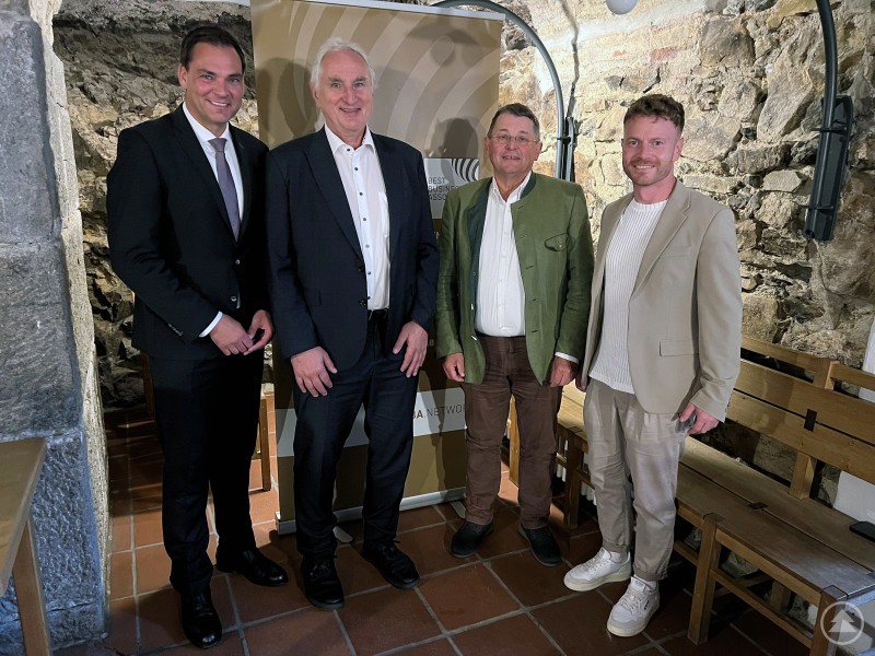 Vier Männer in Anzug und Tracht stehen nebeneinander in einem historischen Gemäuer und lächeln in die Kamera.