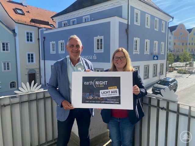 Bürgermeister Günter Streicher und Umweltstadträtin Bianca Scharnböck präsentieren das Plakat zur Teilnahme Schärdings an der „Earth Night“. Ein Mann und eine Frau stehen auf einem Balkon in der Innenstadt von Schärding und halten gemeinsam ein Plakat zur Aktion „Earth Night“ hoch. Im Hintergrund sind historische Gebäude und ein sonniger Himmel zu sehen.