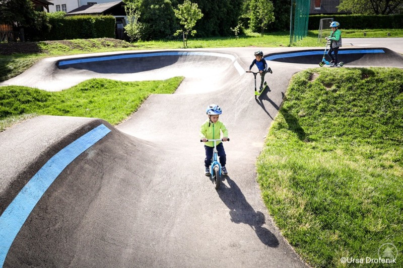 Auf einem Pumptrack können Kinder spielerisch Radfahren, Koordination und Balance trainieren. Drei Kinder fahren mit Laufrad und Tretrollern über einen Pumptrack mit Asphaltwellen und Kurven.