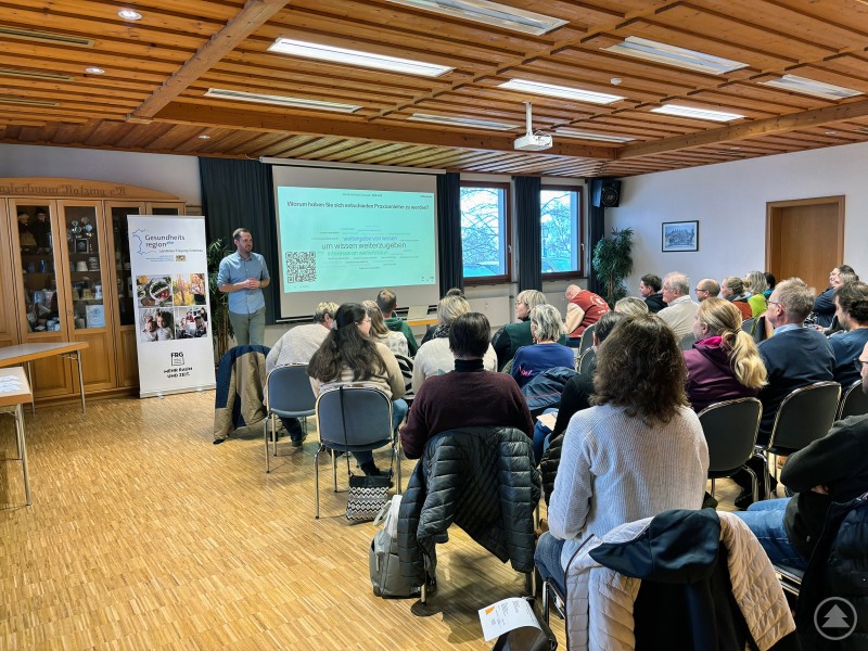 Ein Referent steht vor einer präsentierenden Leinwand und spricht zu einer voll besetzten Zuhörerschaft. Links steht ein Roll-Up der Gesundheitsregionplus Freyung-Grafenau, im Raum sitzen zahlreiche Teilnehmende auf Stuhlreihen.