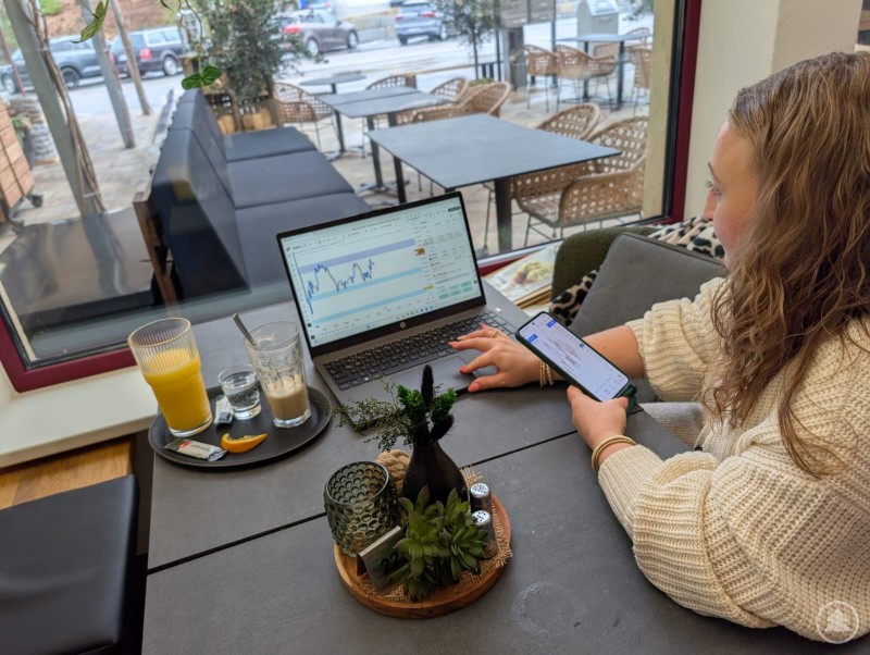 Angelika sitzt in einem Cafe, hat den Blick auf ihren Laptop gerichtet und zugleich ihr Handy mit Kursbewegungen in der Hand. 