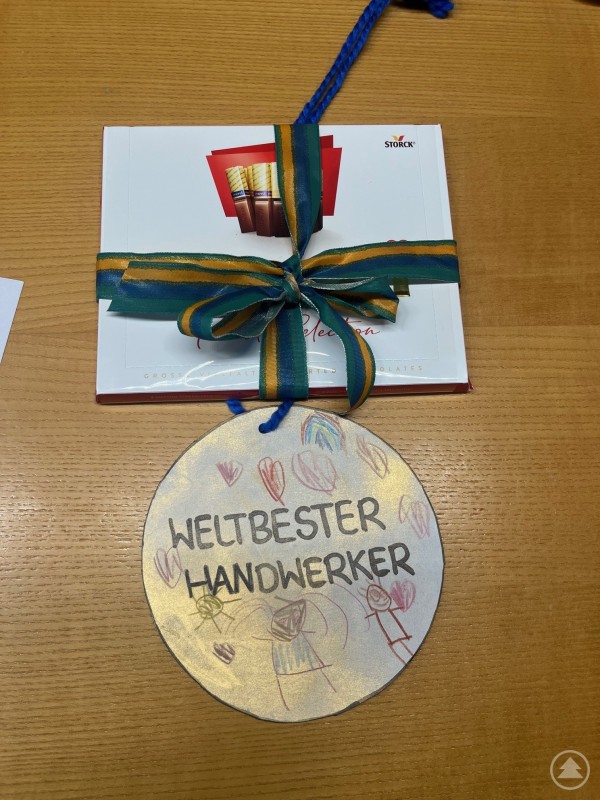 Eine selbst gebastelte Medaille mit der Aufschrift &bdquo;Weltbester Handwerker&ldquo; liegt neben einer Packung Schokolade auf einem Tisch.
