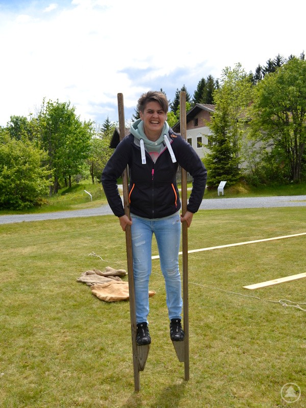 Eine Frau balanciert lachend auf Holzstelzen auf einer Wiese.
