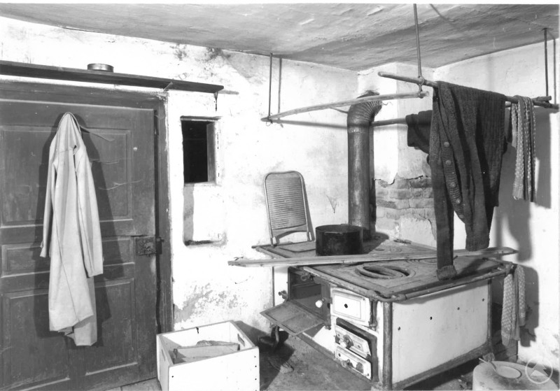 Stube aus dem Tanzerhof im Original, Alter Holzofen, Kleidung oben aufgehängt. Eingangstür mit altem Mantel am Hacken.