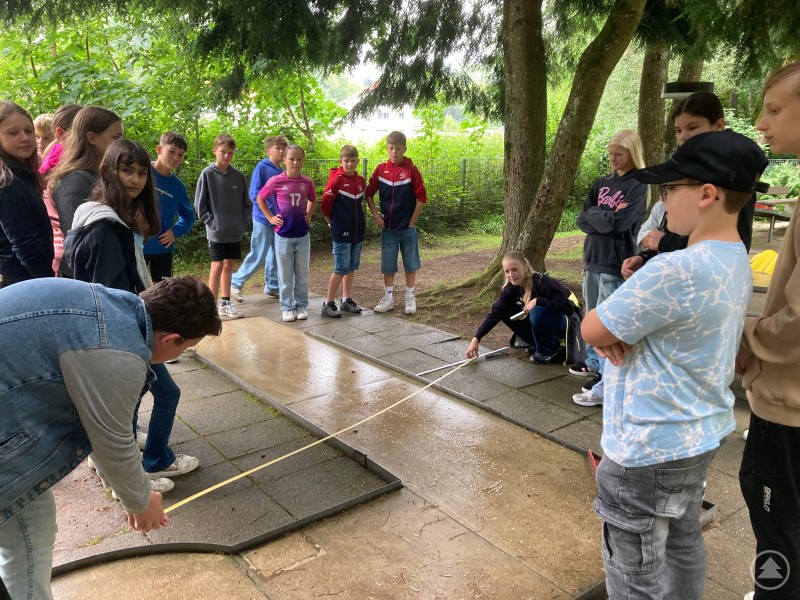 Die Grafenauer Realschüler verlassen sich nicht nur auf ihr Glück, sondern ermitteln zuerst die richtige Laufbahn der Kugel. Eine Gruppe von Schülern beobachtet aufmerksam, wie ein Junge in blauer Kleidung mit einem Minigolfschläger den Ball auf einer nassen Bahn anspielt.
