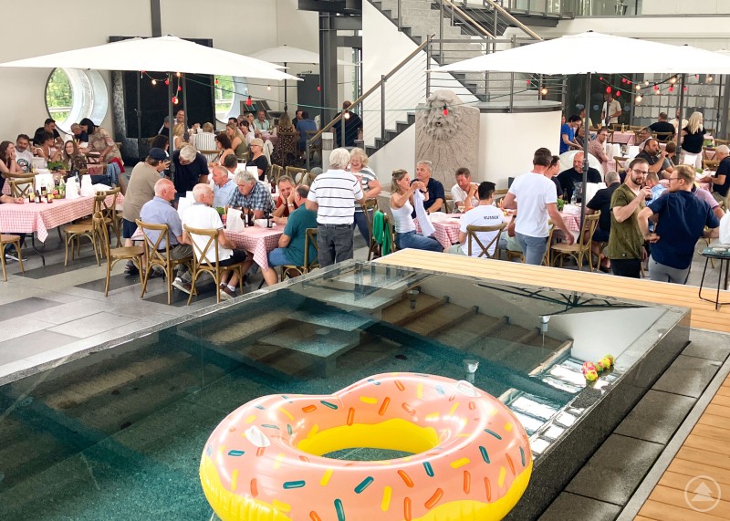 Mediterrane Stimmung, T-Shirt-Aktion und Poolspaß inklusive beim diesjährigen Sommerfest von Kusser Menschen feiern an gedeckten Tischen in einer modernen Ausstellungshalle mit Granitpool
