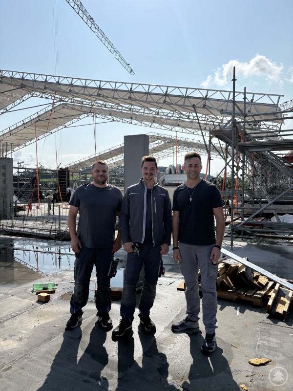 (v. r.) Technischer Leiter Christian Siglmüller, Assistenz Fabian Eibl und Bereichsleiter Patrick Schmid auf der Baustelle des Erweiterungsbaus. Drei Männer in Arbeitskleidung stehen auf einer Baustelle mit Gerüsten, Kränen und provisorischen Dächern.