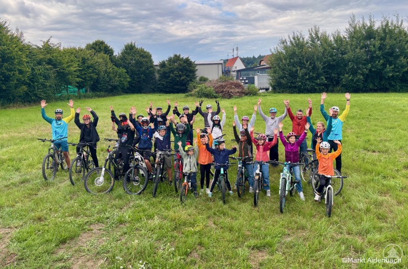 Kinder, Jugendliche und Erwachsene in Aidenbach freuen sich gemeinsam auf den geplanten Bikepark. Eine große Gruppe von Kindern, Jugendlichen und Erwachsenen mit Fahrrädern und Helmen posiert auf einer Wiese und hebt lachend die Hände in die Höhe.