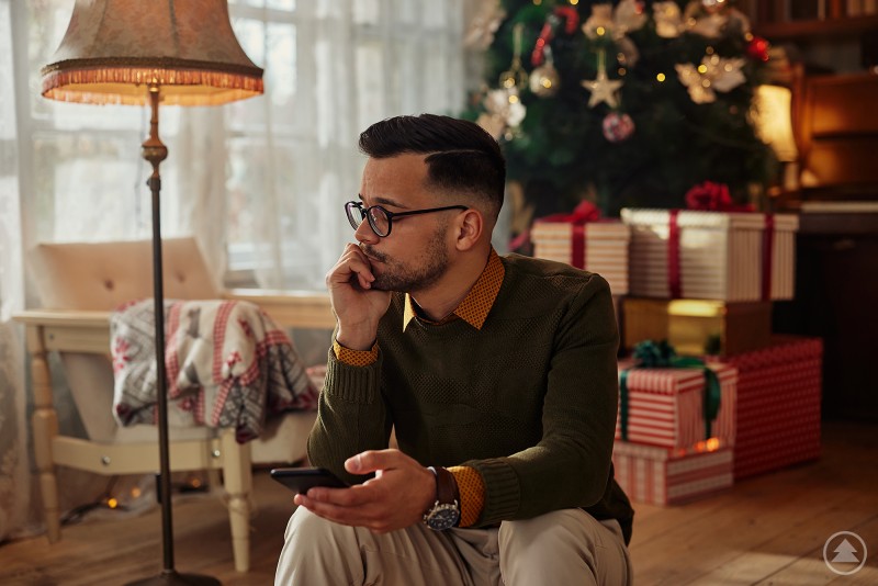 Ein nachdenklich wirkender Mann sitzt in einem weihnachtlich geschmückten Wohnzimmer auf einem Stuhl und hält ein Smartphone in der Hand. Im Hintergrund stehen ein beleuchteter Weihnachtsbaum sowie mehrere verpackte Geschenke. Die Szene vermittelt Ruhe, aber auch innere Anspannung.