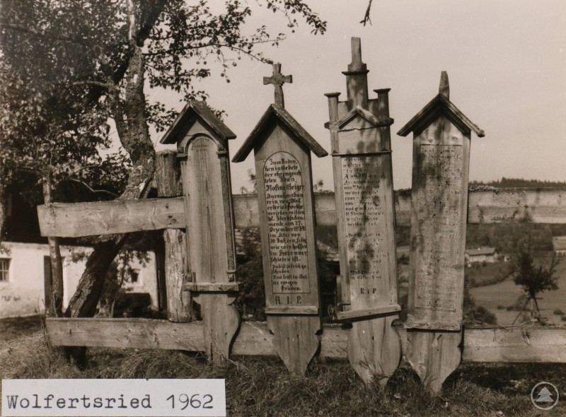 Die Sonderausstellung Jetzt gehts dahi im Freilichtmuseum Finsterau zeigt unter anderem historische Sterbekreuze aus der Region Historische Aufnahme von mehreren hölzernen Sterbekreuzen mit Inschriften, die an einem Zaun befestigt sind. Im Hintergrund sind ein Baum und eine ländliche Landschaft zu sehen.