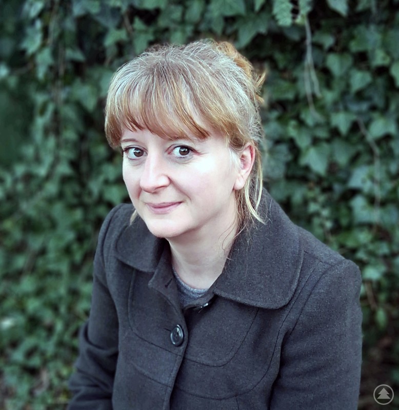 Eine Frau mit blondem Haar, zusammengebundenem Dutt und Pony sitzt im Freien vor einer grünen Efeuhecke. Sie trägt einen dunkelgrauen Mantel und blickt freundlich in die Kamera – ein natürliches, sympathisches Porträt bei Tageslicht.