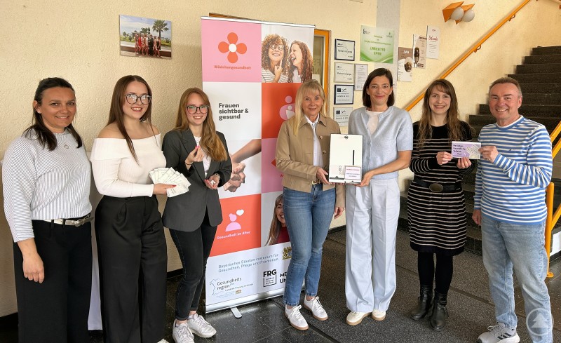 Freuen sich stellvertretend für alle teilnehmenden Schulen über das Gemeinschaftsprojekt (v.r.n.l.): Robert Eberle (Standortleiter Berufliches Schulzentrum Grafenau), Dr. Karin Drachsler (Landratsamt), Evi Blach (Sponsorin der Spender), Elvira Wudy-Engleder (Schulleiterin Berufliches Schulzentrum), Lena Vernay, Nadja Schreiner (beide SMV) und Tanja Müller (Landratsamt). Das Bild zeigt eine Gruppe von sieben Personen, die gemeinsam für ein wichtiges gesundheitliches und gesellschaftliches Thema stehen: kostenfreie Menstruationsprodukte an Schulen. Im Zentrum steht ein Aufsteller mit der Aufschrift „Mädchengesundheit – Frauen sichtbar & gesund“, flankiert von Logos bayerischer Initiativen und Ministerien. Einige der Beteiligten halten Tampons, Binden oder Informationsmaterial in der Hand, eine Frau präsentiert einen weißen Spender für Hygieneartikel. Die Szene wirkt engagiert und kooperativ – ein gelungenes Beispiel für Gesundheitsförderung und Aufklärung in Bildungseinrichtungen, insbesondere im Landkreis Freyung-Grafenau.