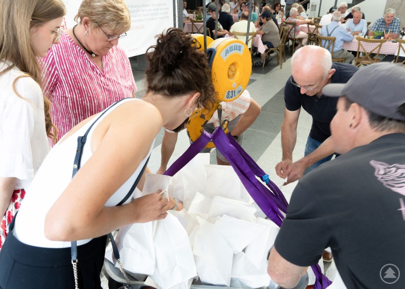 Große Spannung beim Absenken der T-Shirt-Schubkarre und alle sind gespannt, welches Motto auf ihrem Shirt steht. Mehrere Personen beugen sich neugierig über eine Schubkarre, die mit weißen Papiertüten gefüllt ist und an einem Kran befestigt ist.