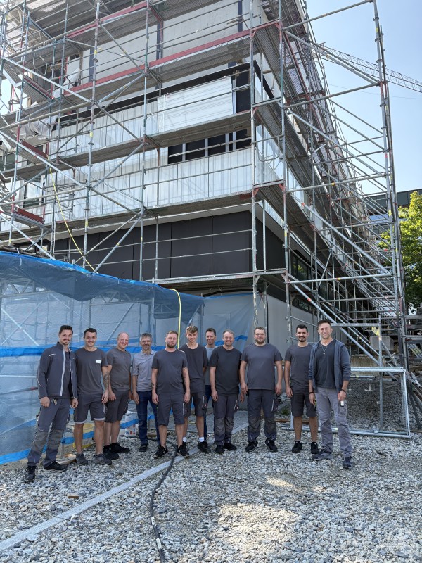 Das gesamte Team der technischen Abteilung am Krankenhaus Freyung. Eine Gruppe von Bauarbeitern und Technikern steht vor einem eingerüsteten Krankenhausgebäude.