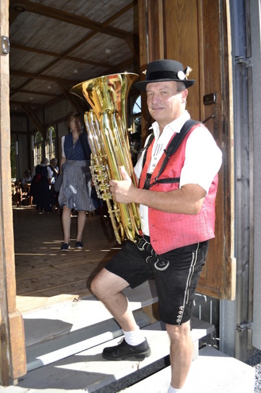 Auftanzgruppe der Ein Mann in traditioneller Tracht mit roter Weste und schwarzem Hut steht mit einer Tuba am Eingang eines Holzgebäudes. Hinter ihm betreten weitere Besucher den Raum.