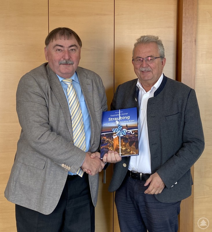 Alfons Lermer (links) wird von Landrat Josef Laumer verabschiedet. Zwei Männer stehen nebeneinander und reichen sich die Hand. Einer von ihnen überreicht dem anderen ein Buch mit blau-weißem Band. Beide lächeln in die Kamera, im Hintergrund ist eine Holzvertäfelung zu sehen.