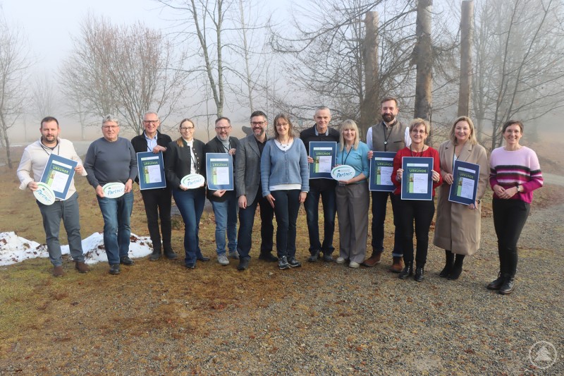 Nationalparkleiterin Ursula Schuster (r.), Alois Grobauer, Vorsitzender des Nationalpark-Partner-Vereins (6.v.l.), und Nationalparkmitarbeiterin Katrin Wachter (7.v.l.) begrüßten die Vertreter der neuen Partner-Betriebe. Eine Gruppe von rund einem Dutzend Personen steht im Freien vor nebliger Waldkulisse. Einige halten Urkunden oder Partnerschilder des Nationalparks in den Händen.