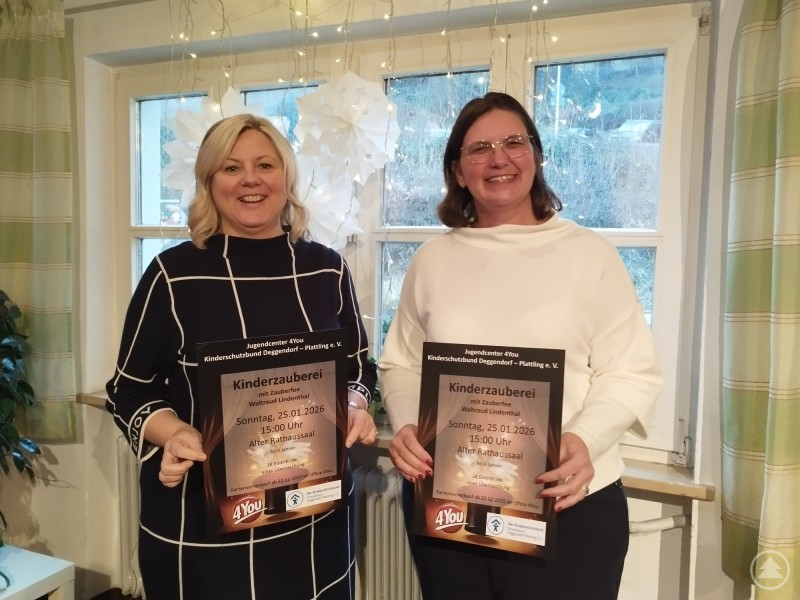 Die Vorsitzende des Kinderschutzbundes Yvonne Pletl-Schäfer (links) und 4You-Leiterin Karin Loibl präsentieren das Plakat für die Kinderzauberei im Alten Rathaus. Zwei Frauen stehen nebeneinander in einem hellen Raum und halten jeweils ein Veranstaltungsplakat in den Händen. Im Hintergrund sind Fenster mit winterlicher Dekoration und Lichterketten zu sehen. Beide lächeln in die Kamera und präsentieren die Ankündigung für eine Kinderveranstaltung.