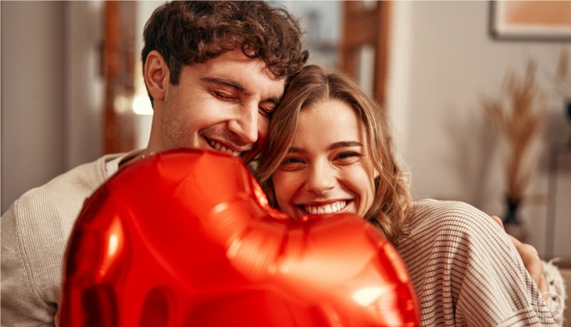 Gemeinsame Zeit schenken macht glücklich – der Valentinstag ist perfekt für kleine Gesten voller Liebe. Ein junges Paar sitzt eng beieinander und lacht glücklich in die Kamera. Im Vordergrund halten sie gemeinsam einen großen, roten Herzluftballon. Die Stimmung wirkt verliebt und herzlich, der Hintergrund ist unscharf.