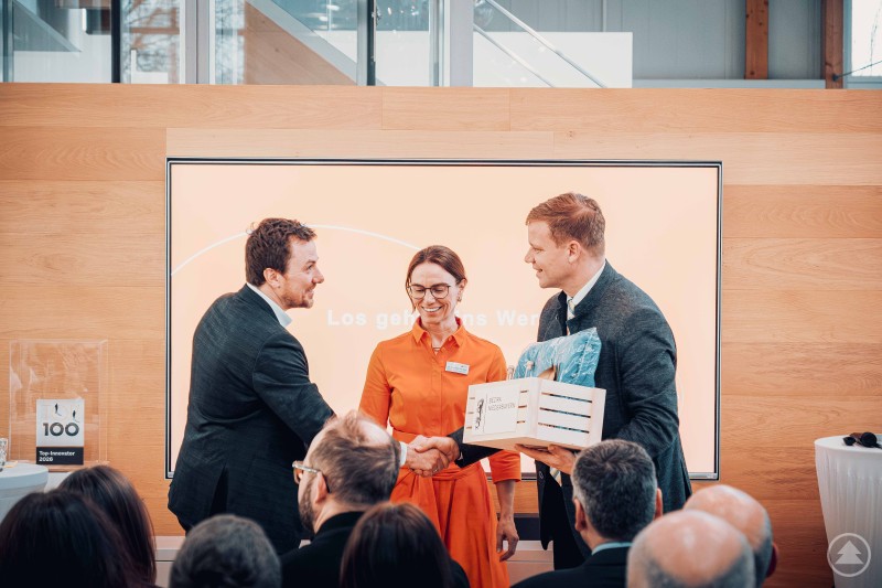 Bezirkstagspräsident Dr. Olaf Heinrich, Vorsitzender des Niederbayern-Forums (rechts), bedankte sich bei Xaver Haas und Dr. Tanja Haas-Lensing für die spannenden Einblicke Drei Personen stehen auf einer Bühne und überreichen eine Auszeichnung vor Publikum in einem modernen Veranstaltungsraum.