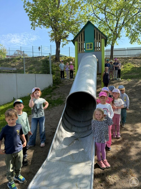 Schönberg ist zum Glück nicht flach. Da passt die Rutsche an den Hang. Mehrere Kindergartenkinder stehen neben einer großen Hangrutsche mit Spielturm auf dem Außengelände des Kindergartens.