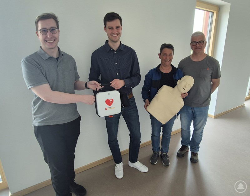 Bei der Übergabe des neuen Defibrillators für das Hallenbad in Bogen: Alexander Oswald (Hallenbad Bogen), Karin Kerscher (Hochbauverwaltung Landratsamt), Simon Geisberger (Leiter Hochbauverwaltung Landratsamt) und Pascal Rogge (Leiter techn. Service Firma HERZkönig, von rechts). Das Bild zeigt vier Personen, die an einer Schulung zur Ersten Hilfe oder zur Defibrillator-Nutzung teilnehmen. Zwei der Personen präsentieren gemeinsam ein AED-Gerät (Automatisierter Externer Defibrillator) mit einem roten Herzsymbol. Eine weitere Person hält eine Reanimationspuppe in Brustgröße – typisch für praktische Übungseinheiten zur Herzdruckmassage. Die Gruppe wirkt gut gelaunt und engagiert, der Hintergrund ist ein heller Schulungsraum. Die Szene vermittelt den wichtigen gesellschaftlichen Beitrag von Wiederbelebungstrainings zur Lebensrettung im Notfall.
