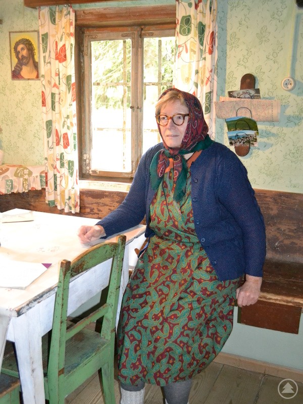 Die letzte Bewohnerin im Sachl – Lina Graf alias Marianne Moosbauer – erzählt vom Leben in den 60er Jahren. Frau im traditionellen Kleid und Kopftuch sitzt in rustikal eingerichteter Bauernküche.