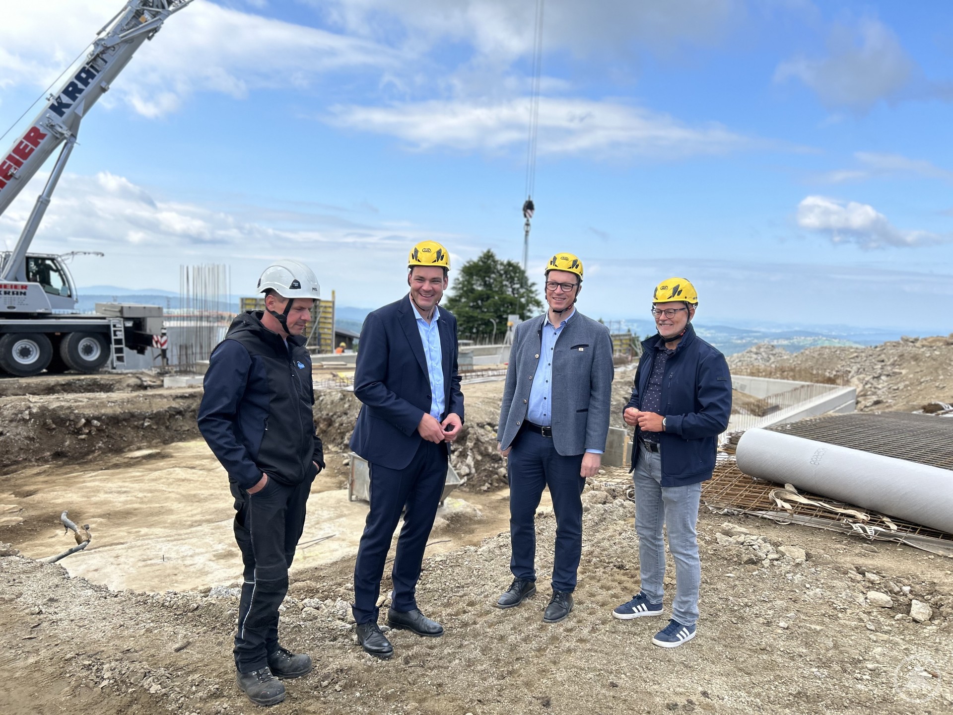 Vier Männer mit gelben Helmen stehen auf einer hochgelegenen Baustelle mit Blick über die hügelige Landschaft. Im Hintergrund ist ein Kran zu sehen, der an einem Seil ein Bauteil hebt.