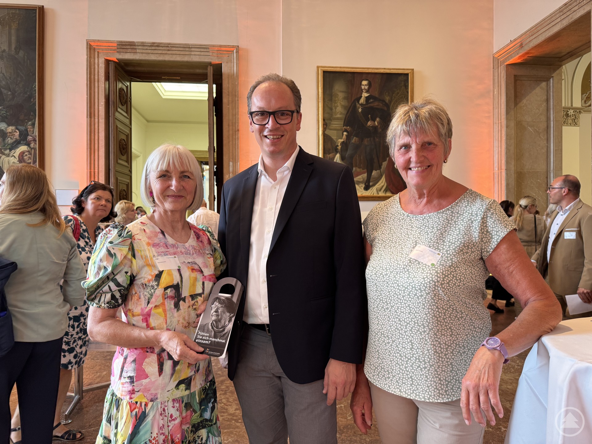 Drei Personen stehen in einem festlichen Saal mit historischen Gemälden an der Wand. Zwei Frauen flankieren einen Mann in Anzug. Die Frau links hält eine Broschüre oder eine Informationsmappe in der Hand.